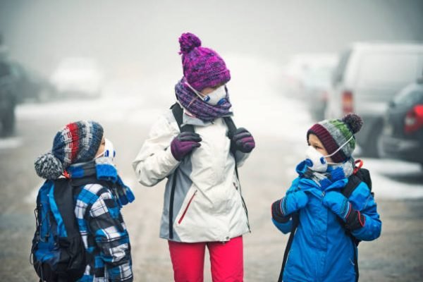 Three kids wearing pollution masks and backpacks are walking to school in the smog. The air is thick with mist and smog.